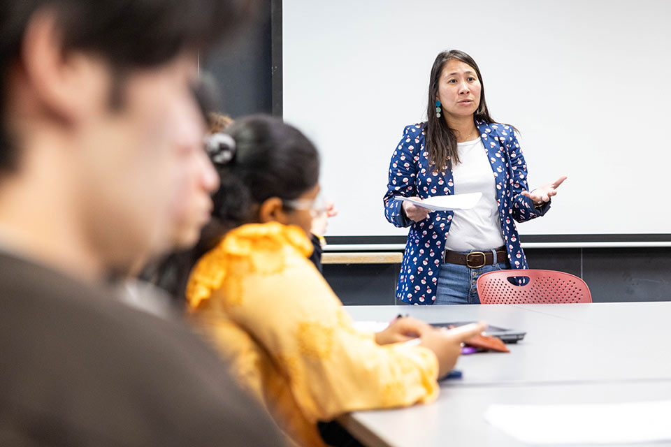 A professor stands in front of a whiteboard in a classroom speaking to students