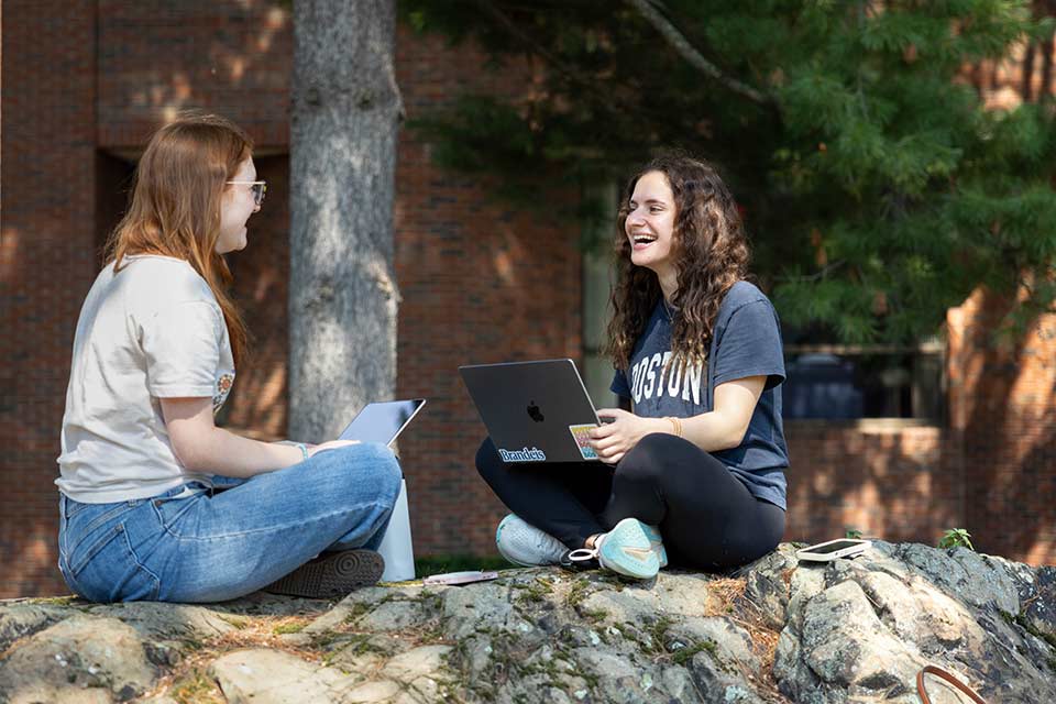 Two students sit on a rock on the Brandeis campus smiling and working on their laptops.