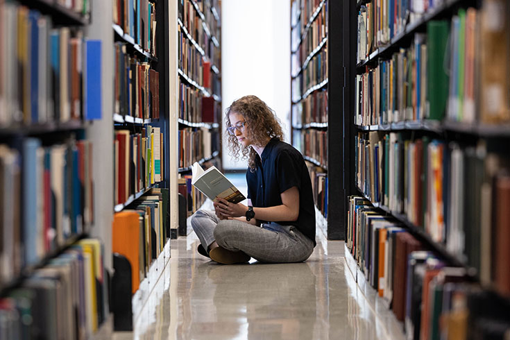 A student sitting on the floor between book shelves in the library reading a book