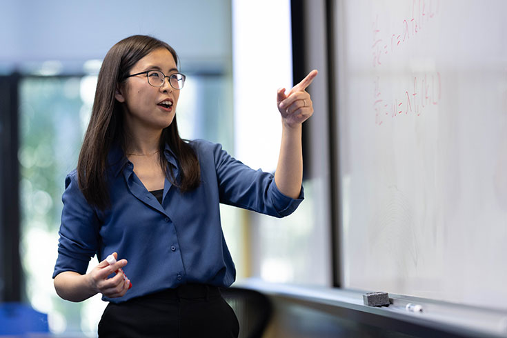A professor points to a whiteboard while teaching a class. 