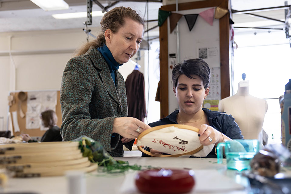 A student embroiders cloth as a professor looks on