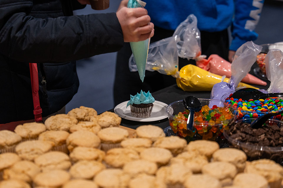 Image shows cupcakes and cupcake decorating supplies on a table. A hand can be seen adding frosting to a cupcake.