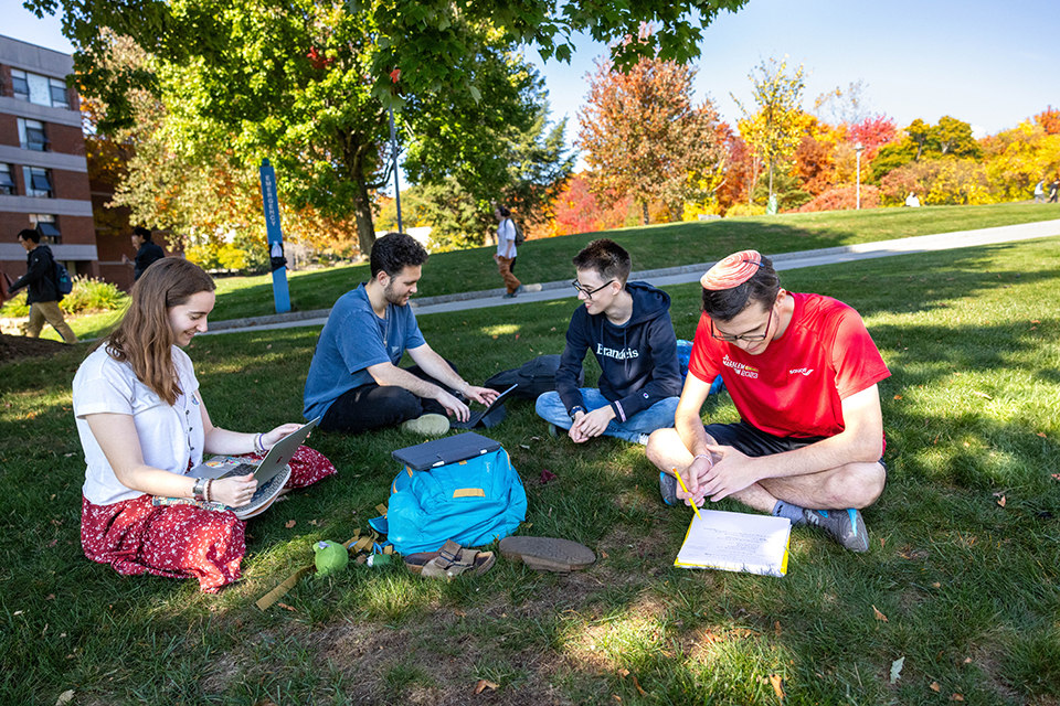 Students sit in a circle on the ground outside doing schoolwork with laptops and notebooks.