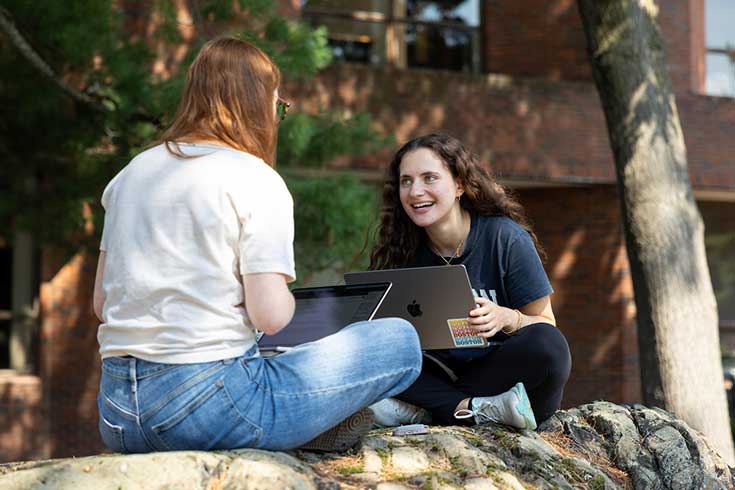 Two students work on laptops outdoors on Brandeis campus.