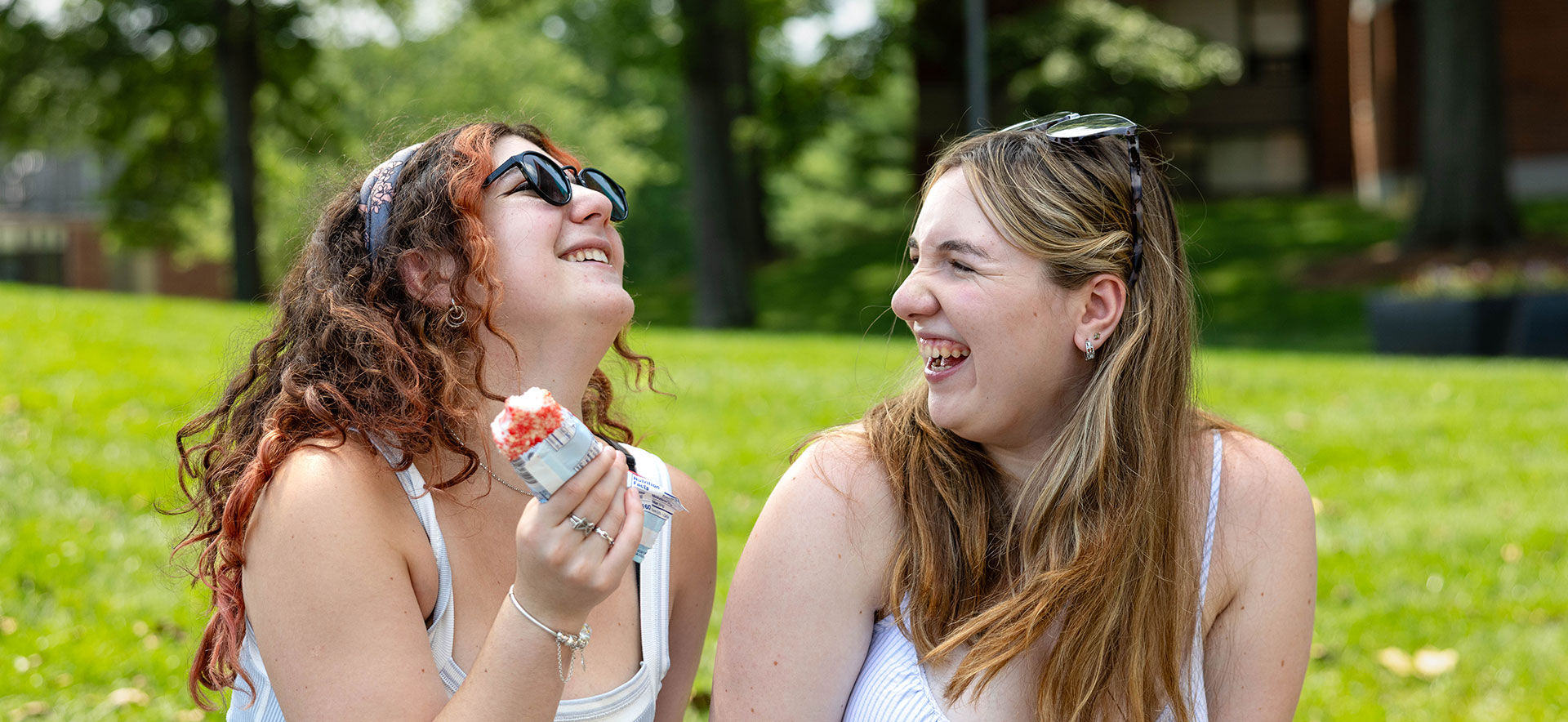 Two students outside on the Brandeis campus laughing and eating ice cream.
