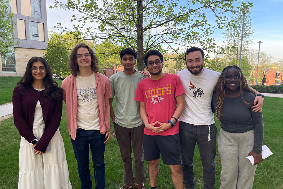 6 students of various ethnic backgrounds standing outside on grass smiling into the camera with a spring tree behind them