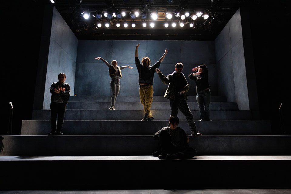 Student actors dance together on a dimly lit stage.