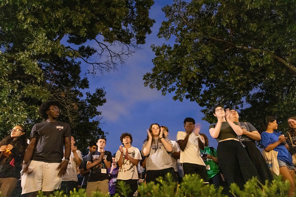 Students standing outside at dusk; some clapping; at the annual Light the Night Orientation event.