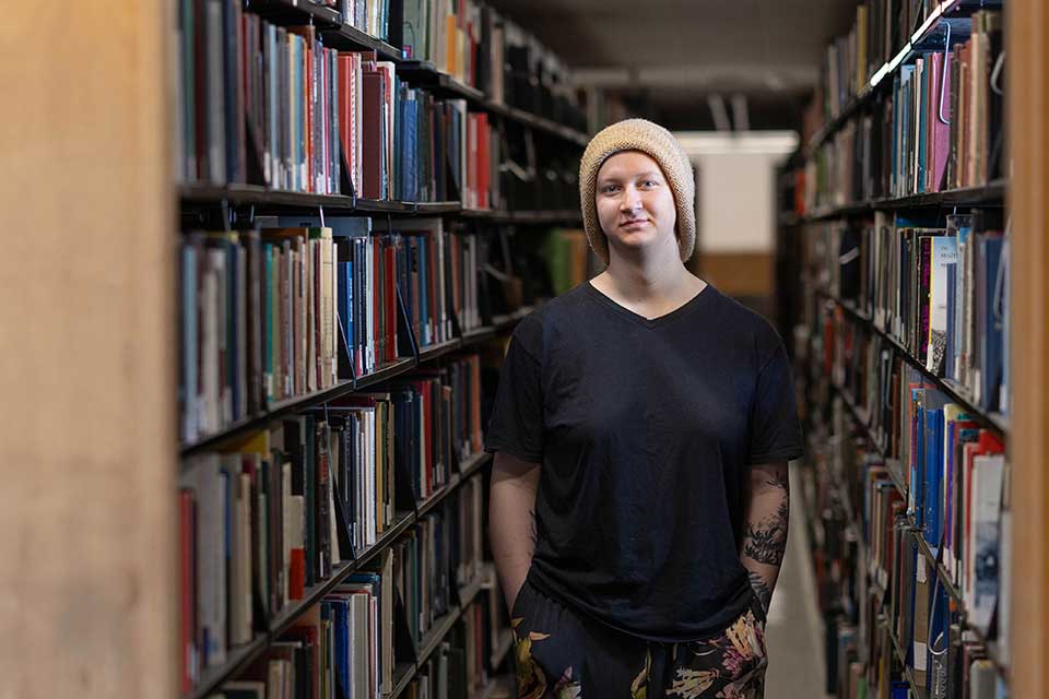 A student stands between shelves of books in the library.