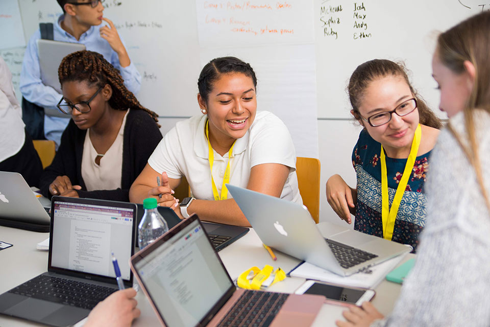 Students wearing lanyards and working on laptops smile while talking together