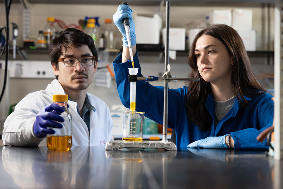 Two graduate students conduct research with a pipette and beakers in a lab.