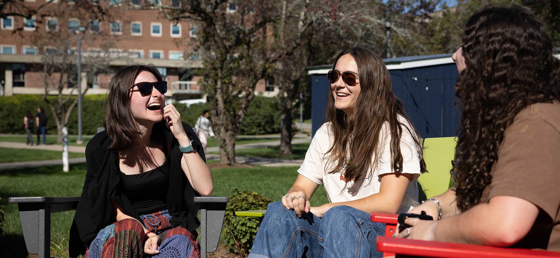 Students wearing sunglasses laugh while sitting outside in the sunshine. 
