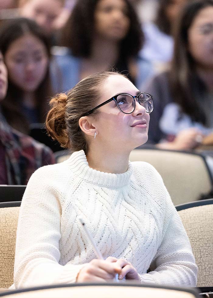 A student sits in a lecture hall looking up at a presentation.
