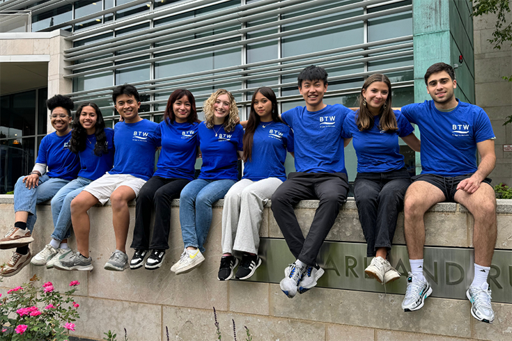 Nine students sit on a wall wearing matching blue BTW shirts.